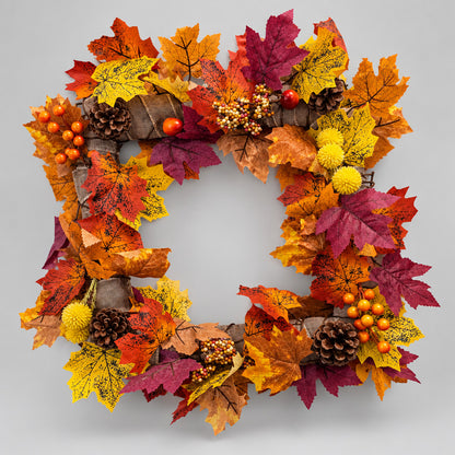An autumn-themed decorative wreath made of faux maple leaves in orange, yellow, red, and purple, with pinecones, small orange berries, and yellow pom-poms, hanging on a plain light gray wall.